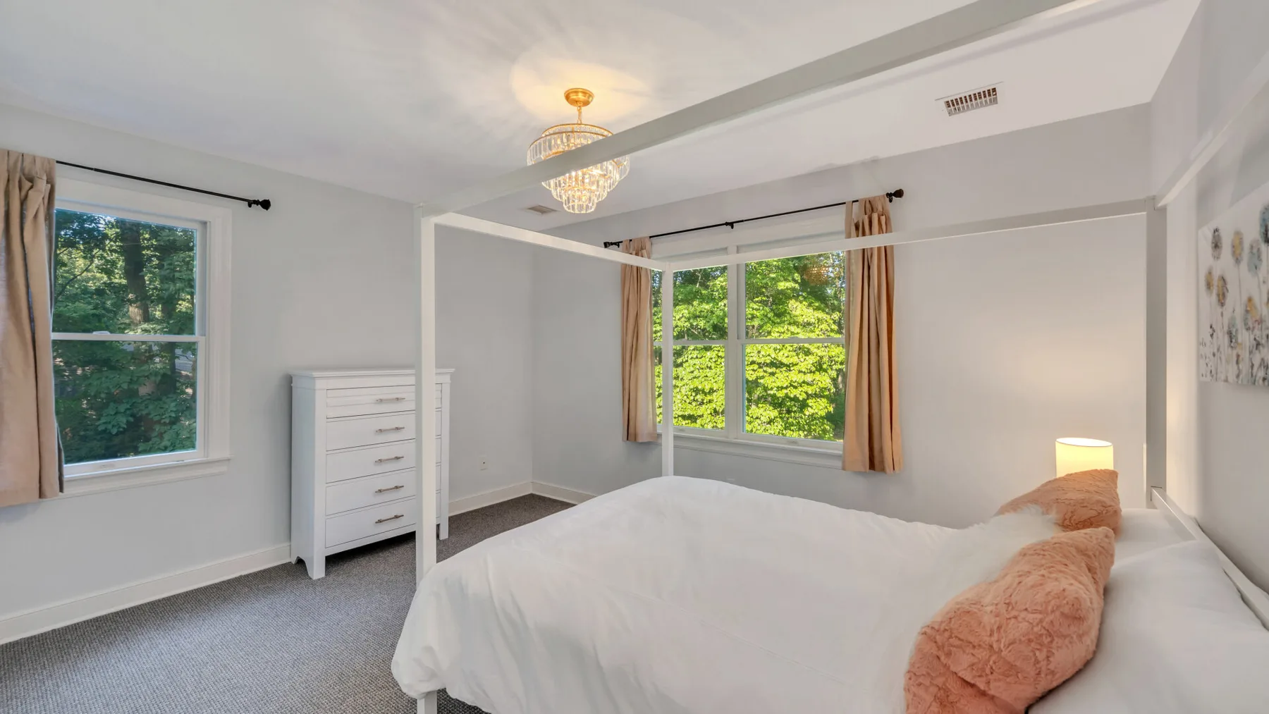 A bright bedroom with a bed, dresser, and windows overlooking green trees.