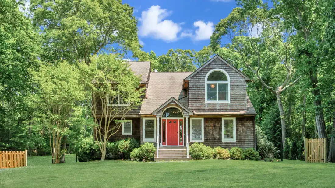 A shingled house with a red front door stands amid trees and a broad lawn.