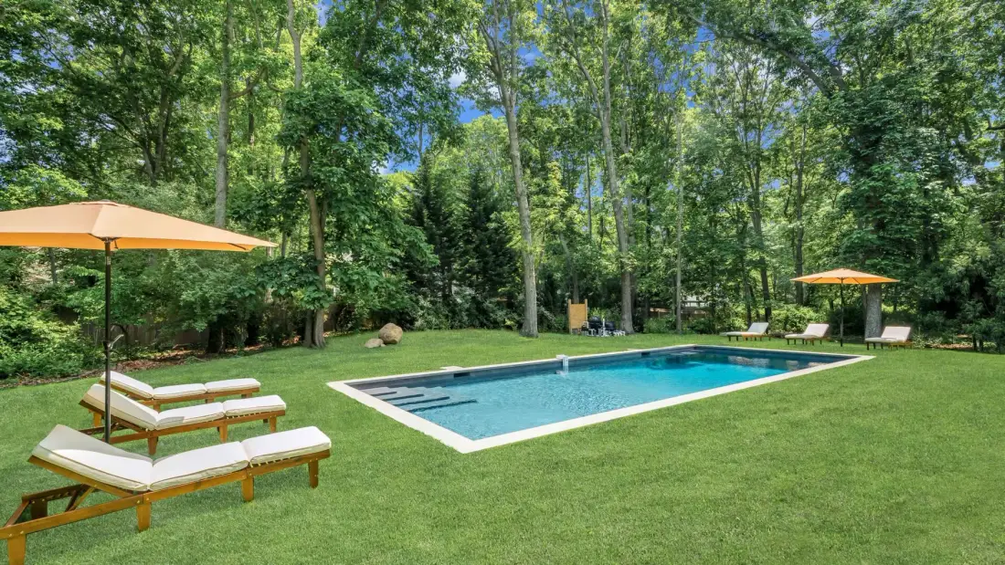 A swimming pool sits in a grassy lawn surrounded by trees and lounge seating.