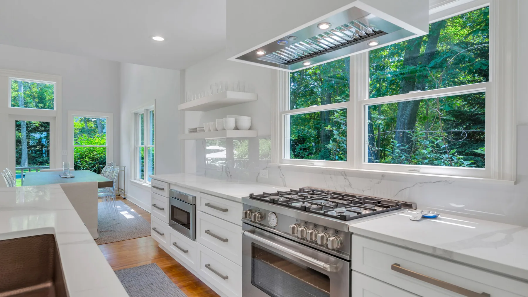 A bright kitchen with white cabinetry, a large range, and wide windows overlooking trees.