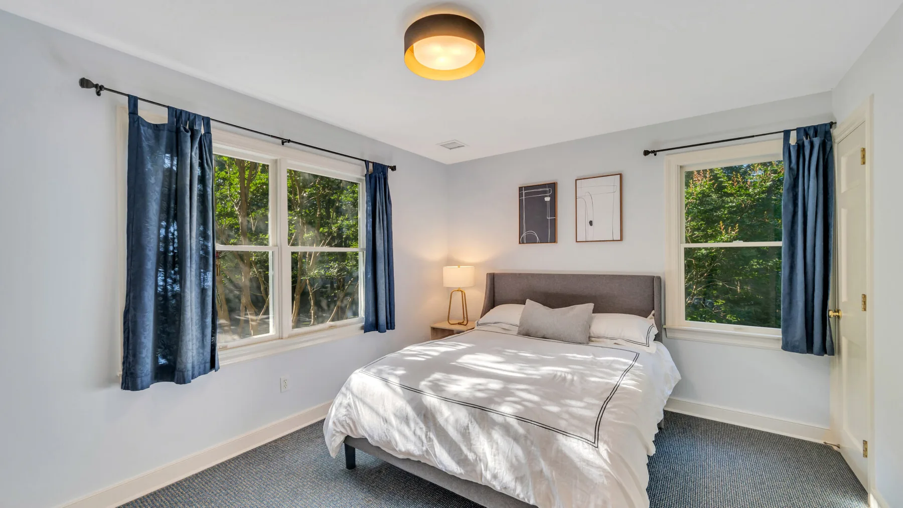 A bedroom with a bed between two windows framed by blue curtains.