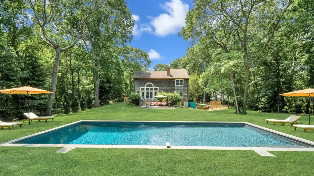 A swimming pool sits in a grassy yard in front of a house surrounded by trees.