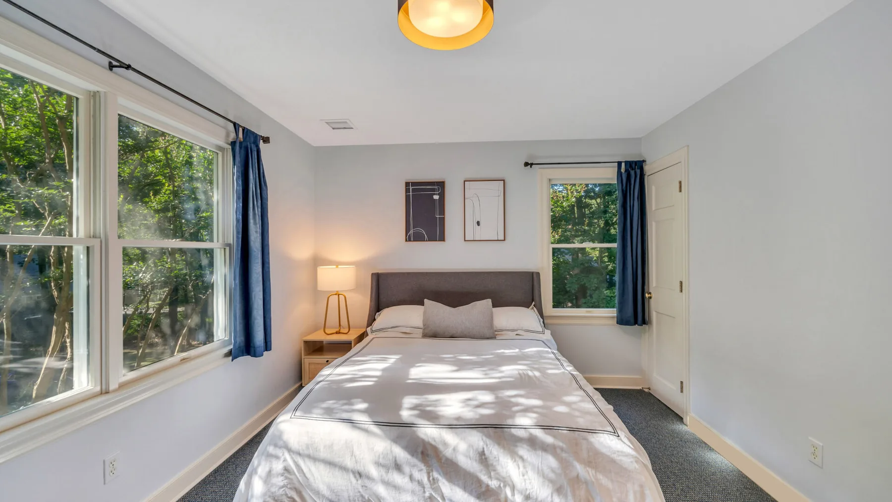 A bedroom with a neatly made bed between large windows and a doorway.