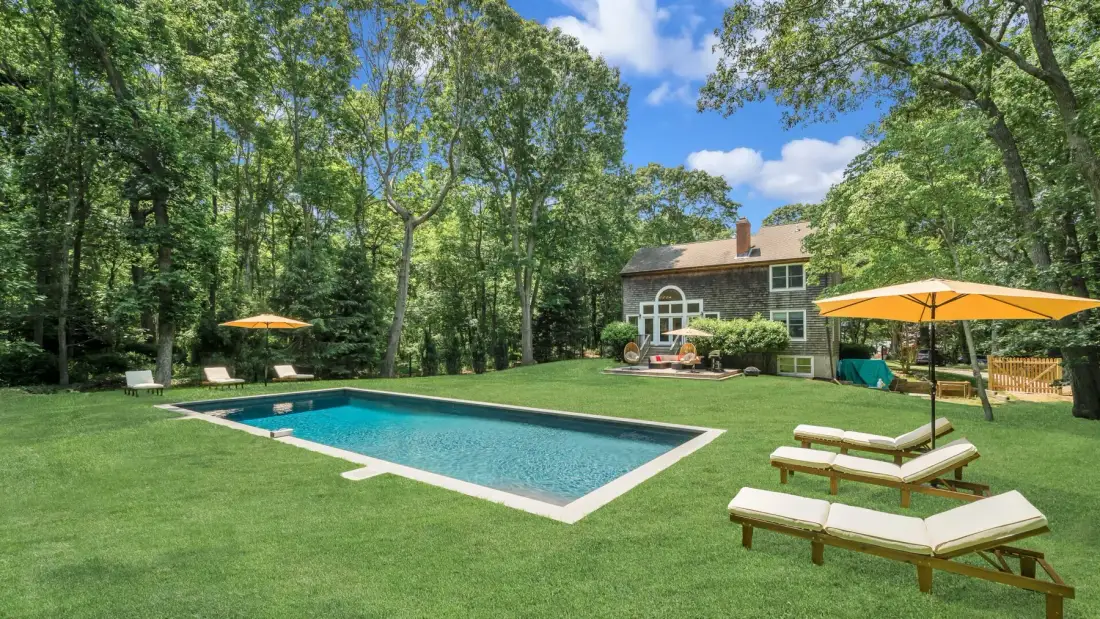 A backyard with a swimming pool, lawn, and trees surrounding a house.