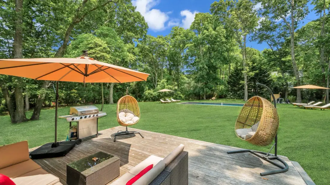 An outdoor deck overlooks a grassy yard, with lounge seating and a large orange umbrella.