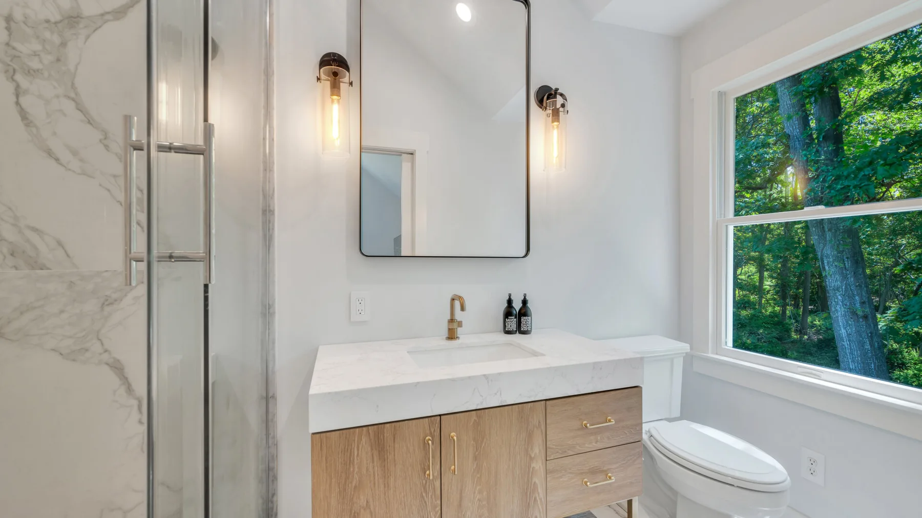 A bright bathroom with a vanity, mirror, and window overlooking trees.
