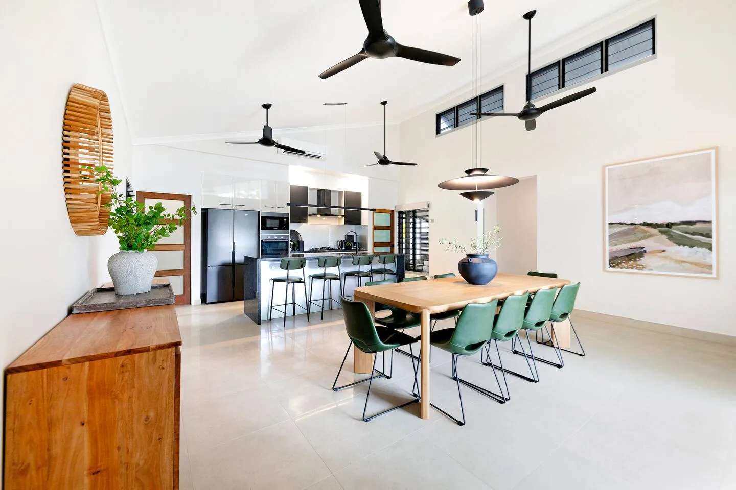 A bright dining area features a long table with green seating beside an open kitchen.