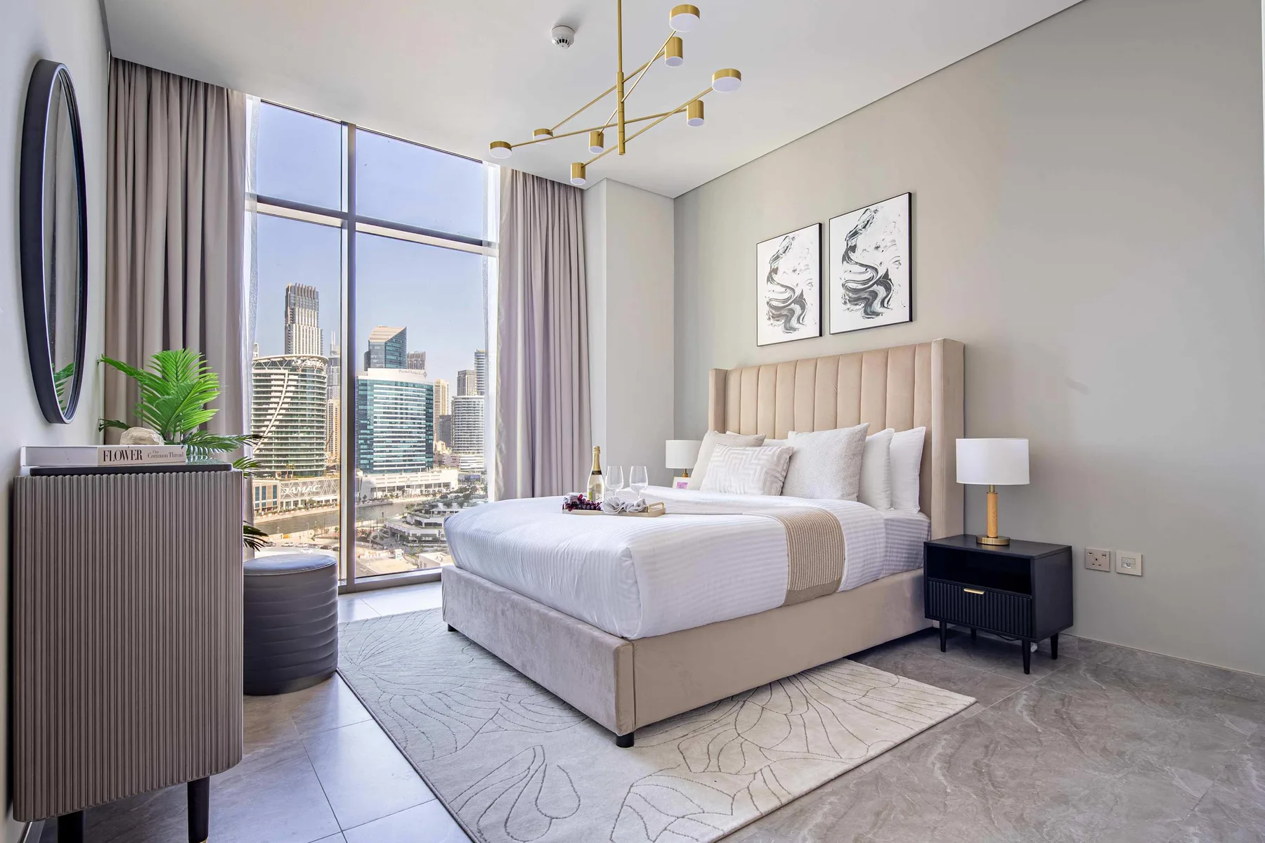 A bright bedroom with a bed beside a floor-to-ceiling window overlooking city buildings.