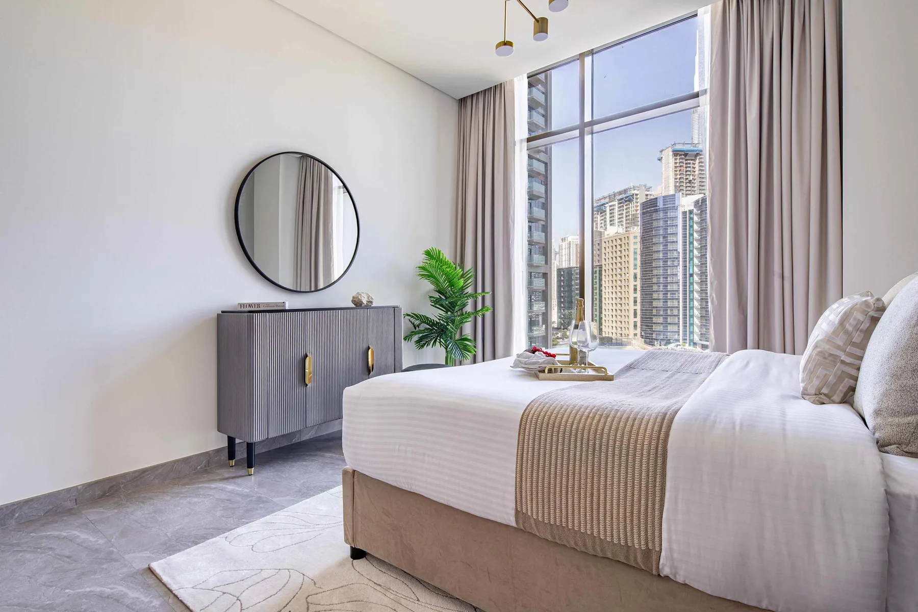A bright bedroom with a bed beside large windows overlooking city buildings.