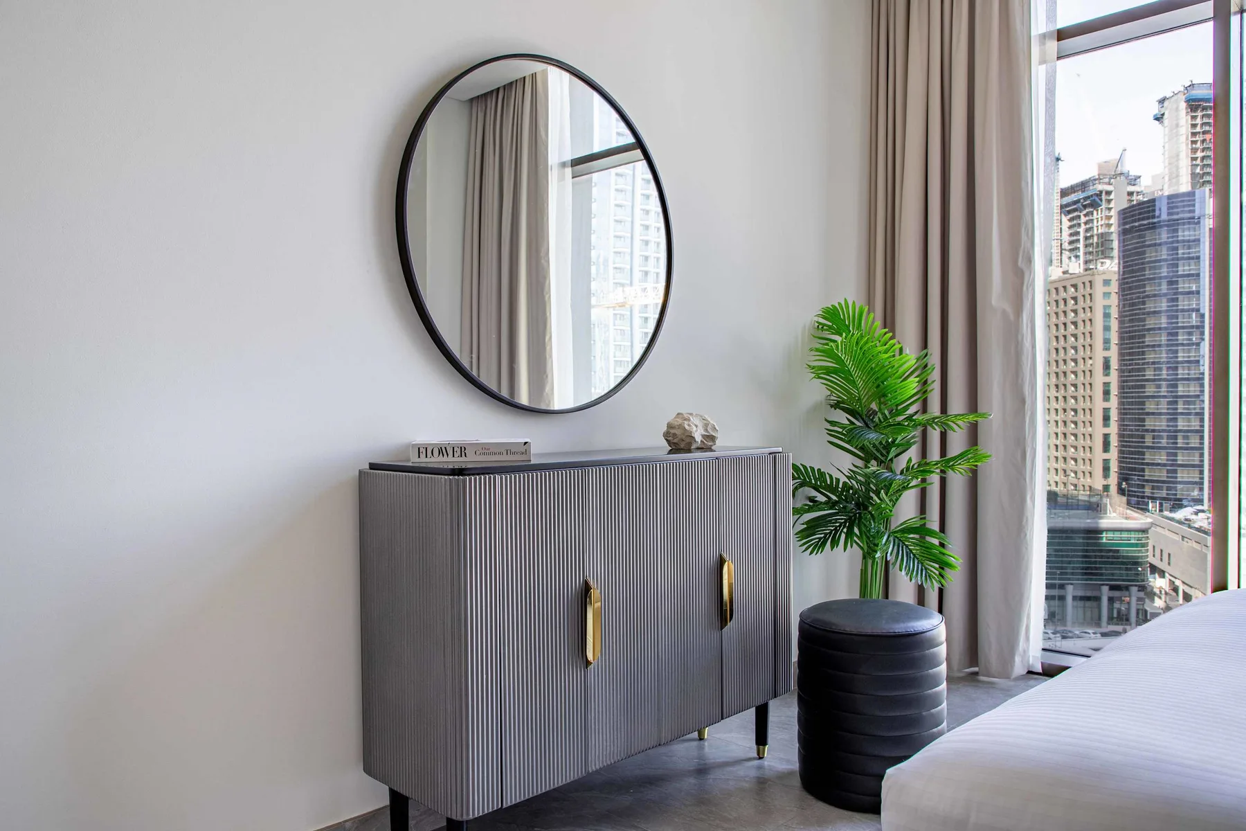 A bedroom corner with a ribbed cabinet, round mirror, and potted plant by the window.
