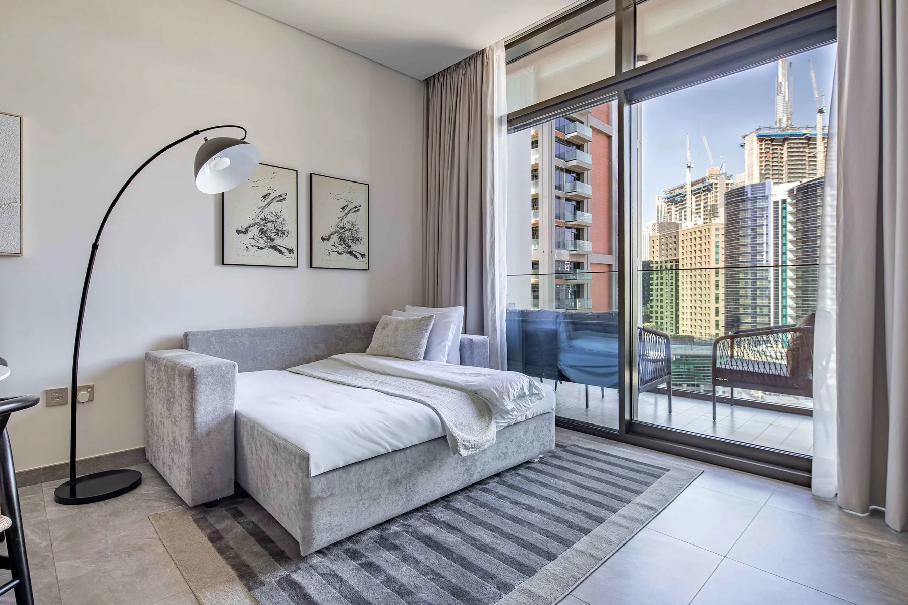 A bedroom with a bed beside floor-to-ceiling windows overlooking nearby buildings.