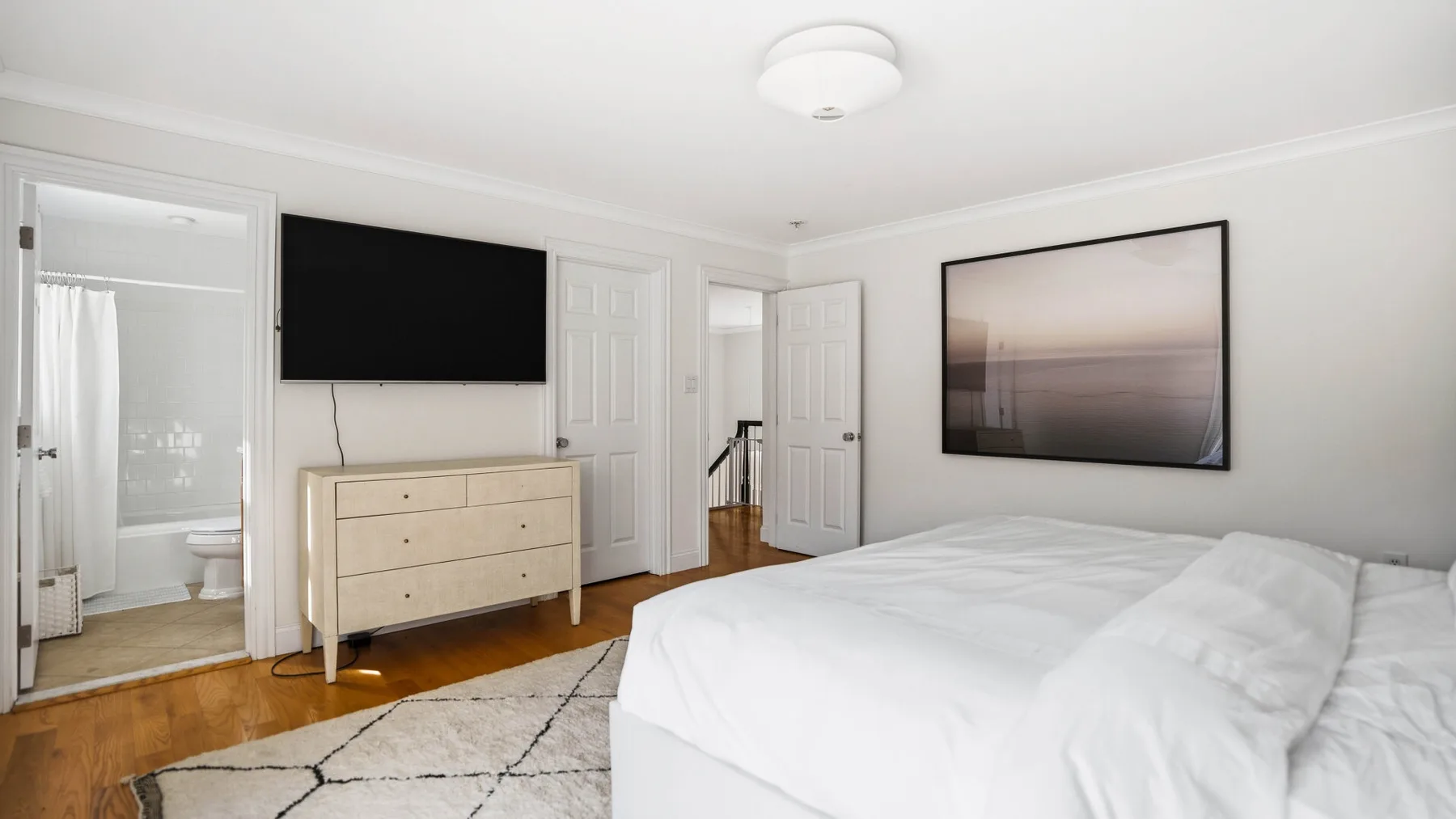 Un dormitorio luminoso con ropa de cama blanca, una cómoda y arte enmarcado.