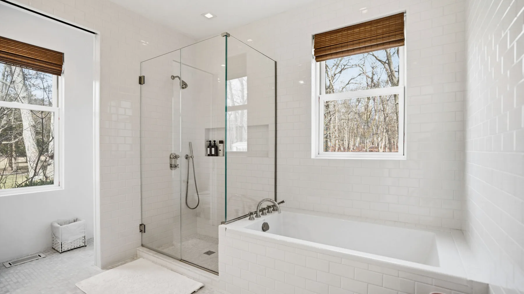 Un baño luminoso con una ducha de vidrio y una bañera junto a dos ventanas.