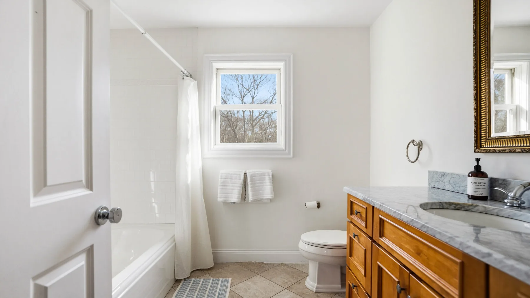 Un baño luminoso con bañera, inodoro y tocador de madera bajo una ventana.