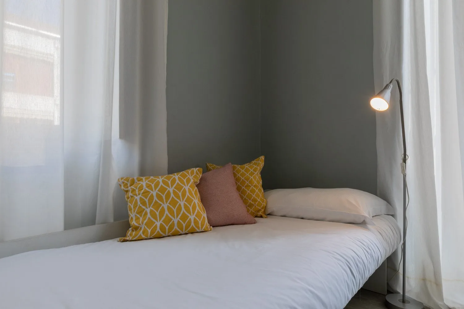 Una cama bien tendida con cojines estampados está junto a una lámpara de pie encendida.