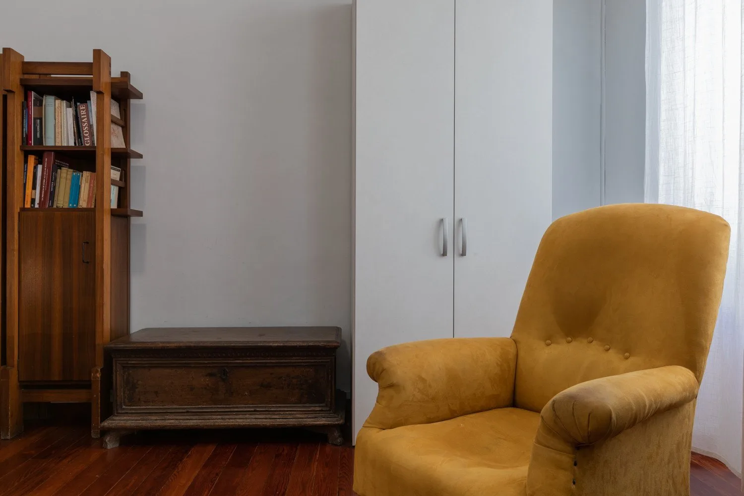 Un sillón color mostaza está junto a una estantería y un baúl de madera en una habitación.