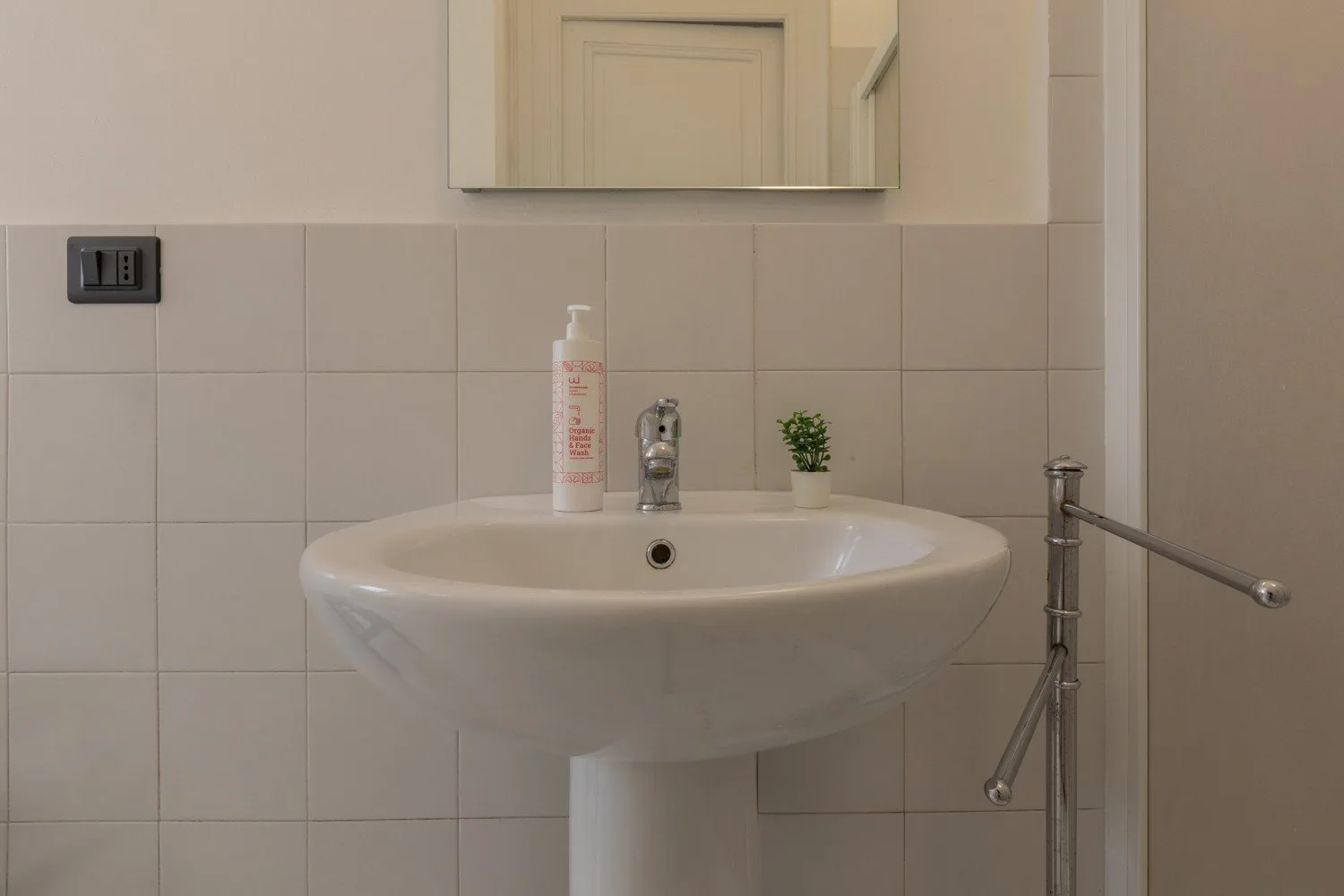 Un lavabo de baño está bajo un espejo, con paredes de azulejos y una planta pequeña.