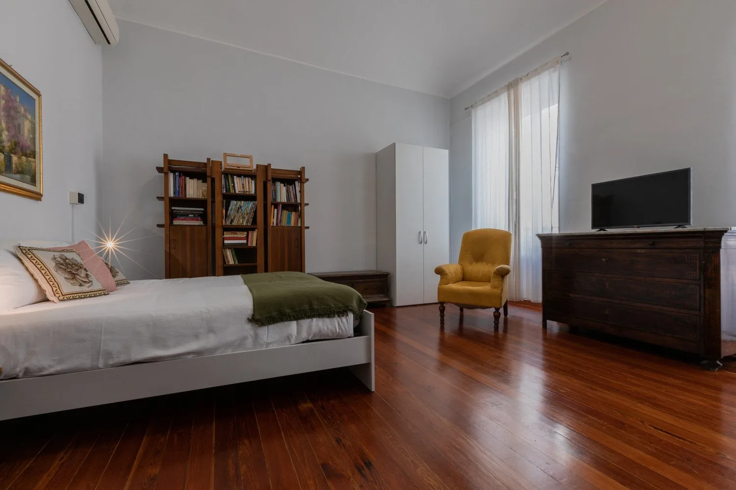Un dormitorio con cama, estantería, sillón y televisor.