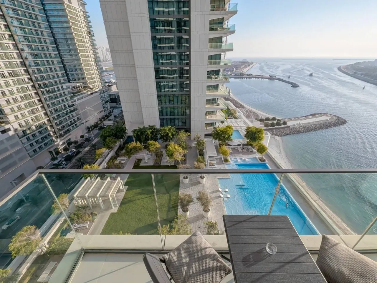 A high-rise balcony overlooks a swimming pool, landscaped grounds, and a waterfront.