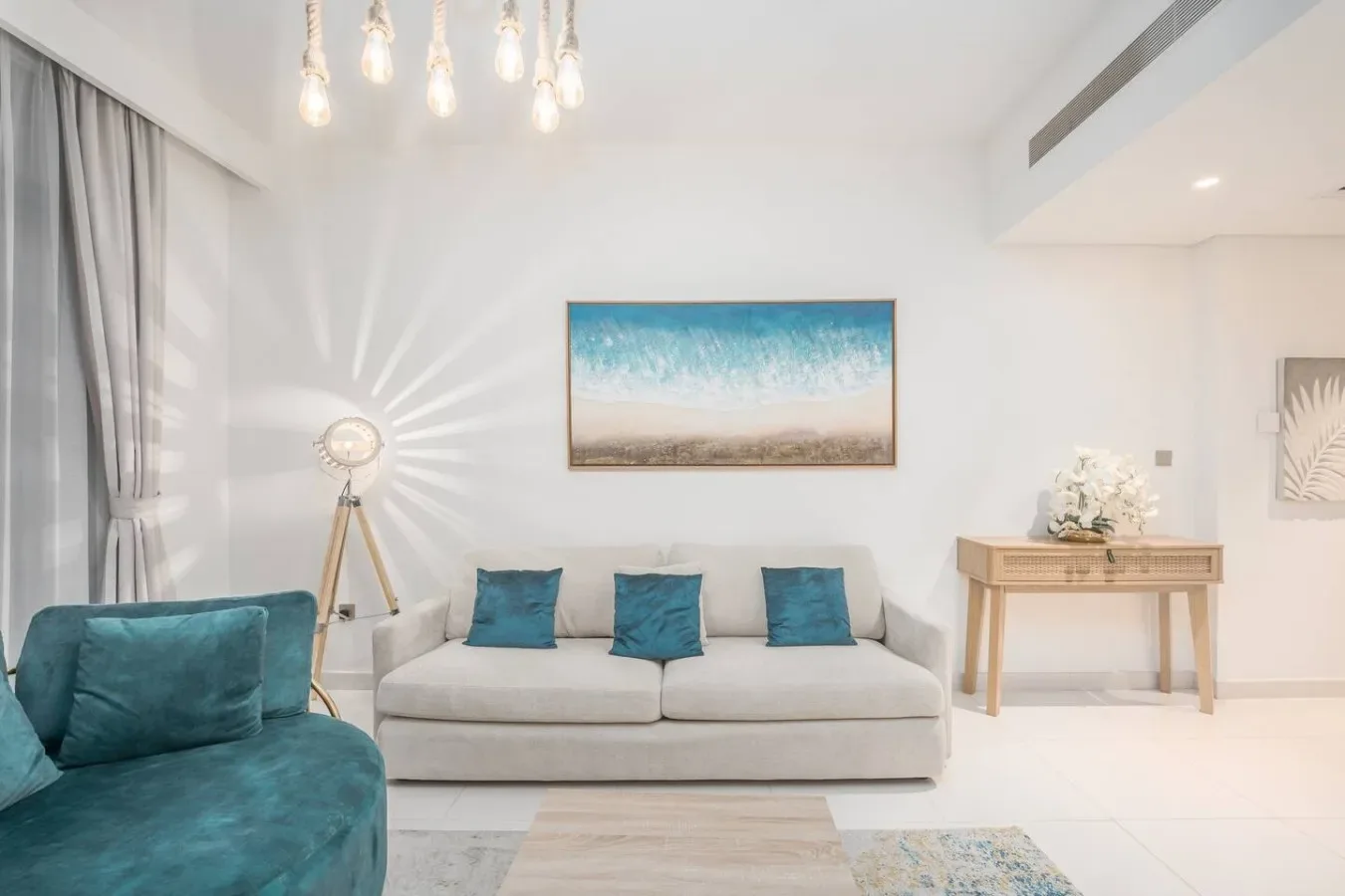Una sala luminosa tiene un sofá claro, asientos color turquesa y arte costero.