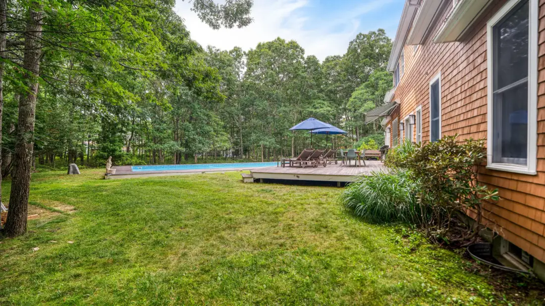 Un patio con césped junto a una casa se abre a una piscina y una zona de estar con sombra.