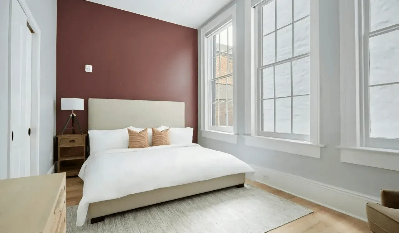 Dormitorio con una cama junto a una pared rojo oscuro y ventanas altas.