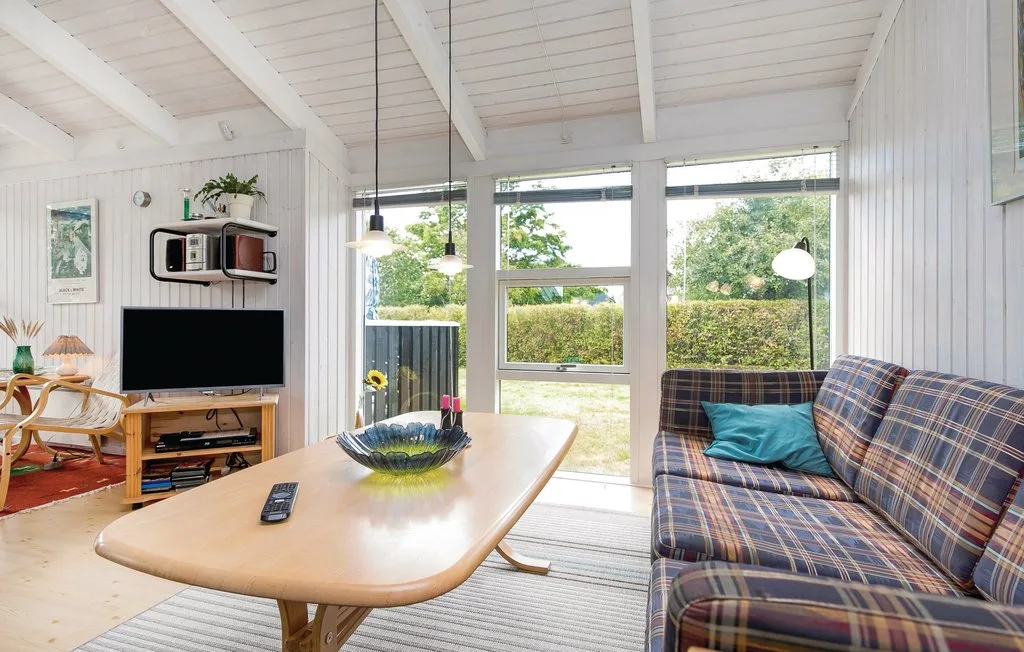 Una sala luminosa con un sofá de cuadros, un televisor y grandes ventanas.