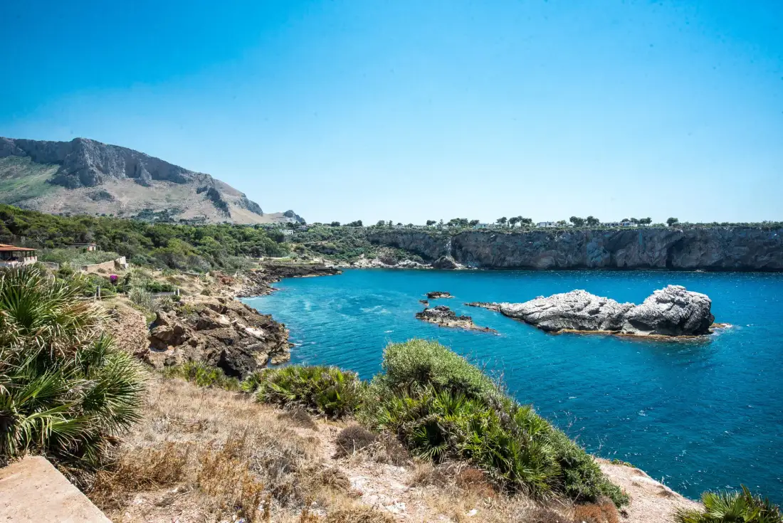 A coastal cove with clear blue water, rocky shores, and low green vegetation.