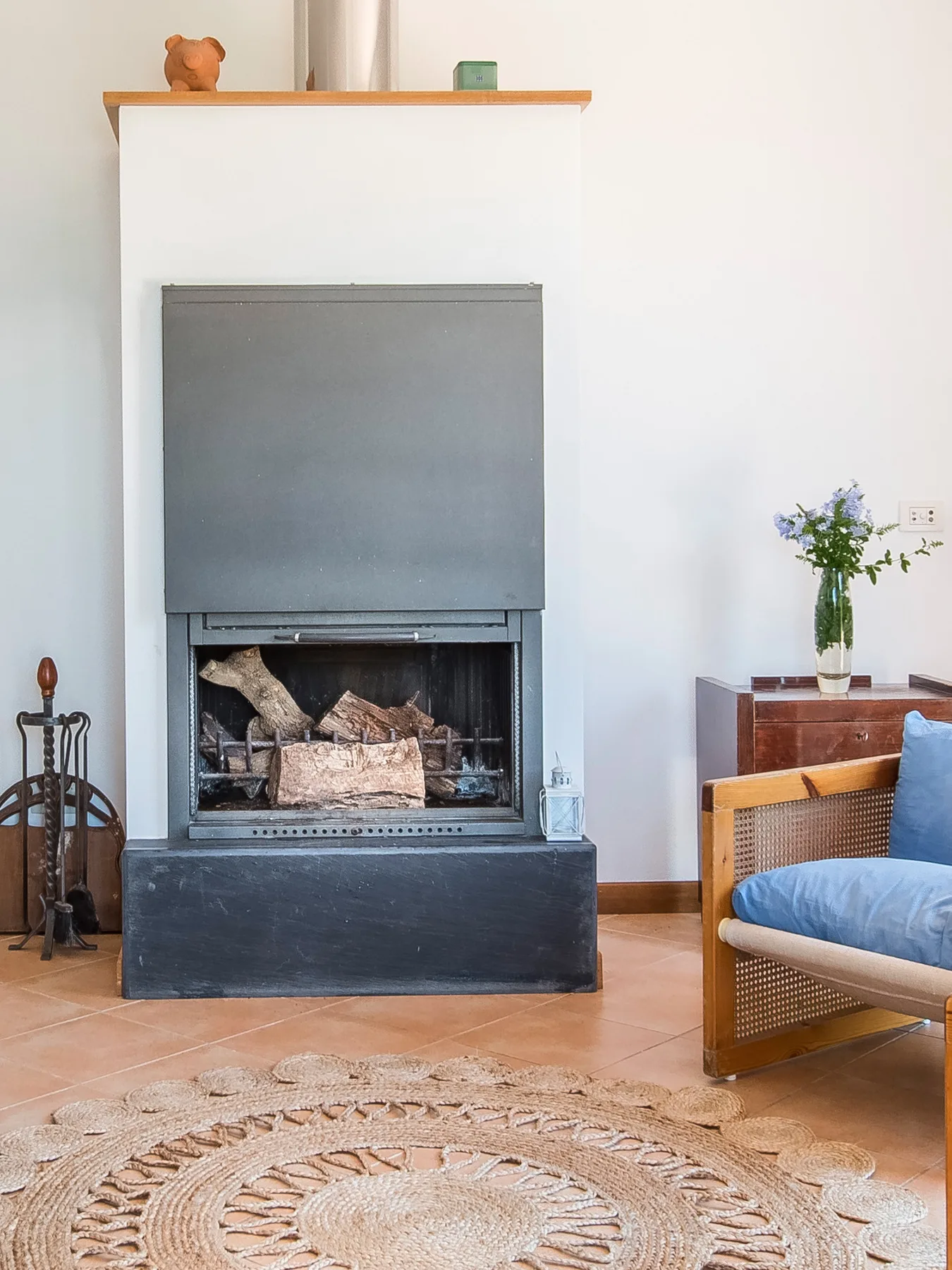 A living room fireplace with stacked logs, beside a chair and round woven rug.