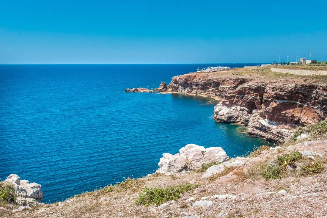 A rocky coastline borders clear blue water beneath a bright sky.