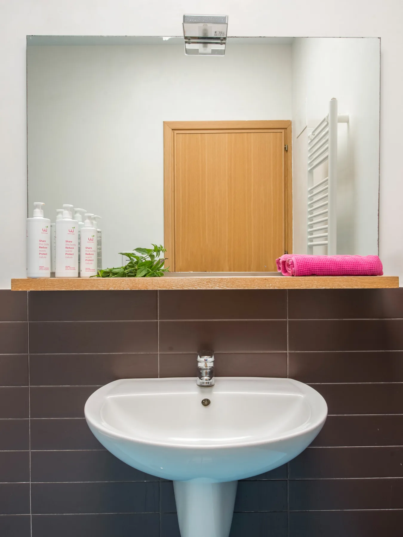 A bathroom sink sits beneath a mirror, with toiletries and a folded pink towel on the ledge.