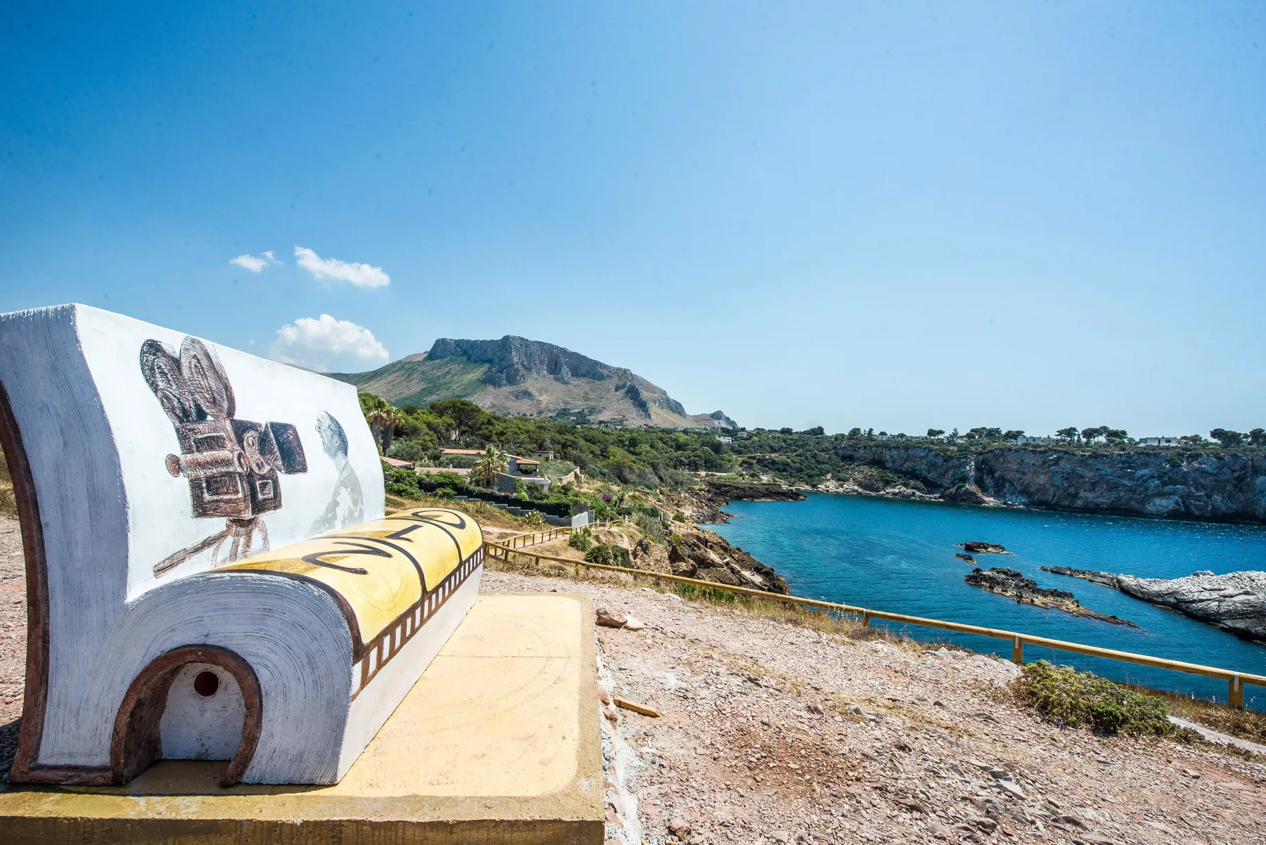 A coastal viewpoint overlooks a blue bay, with a sculpted bench in the foreground.