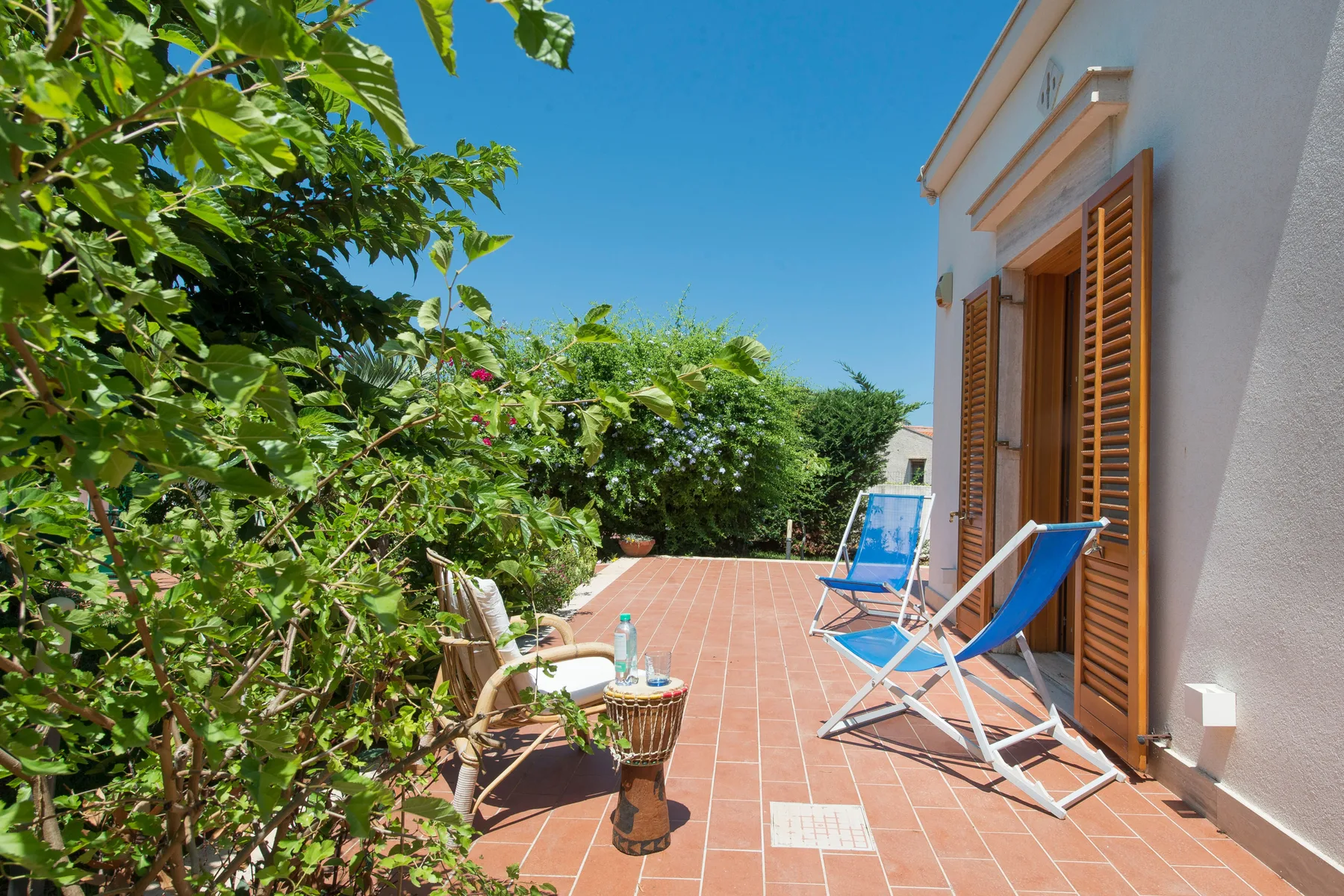 A sunny tiled patio beside a building, with outdoor seating and leafy plants.