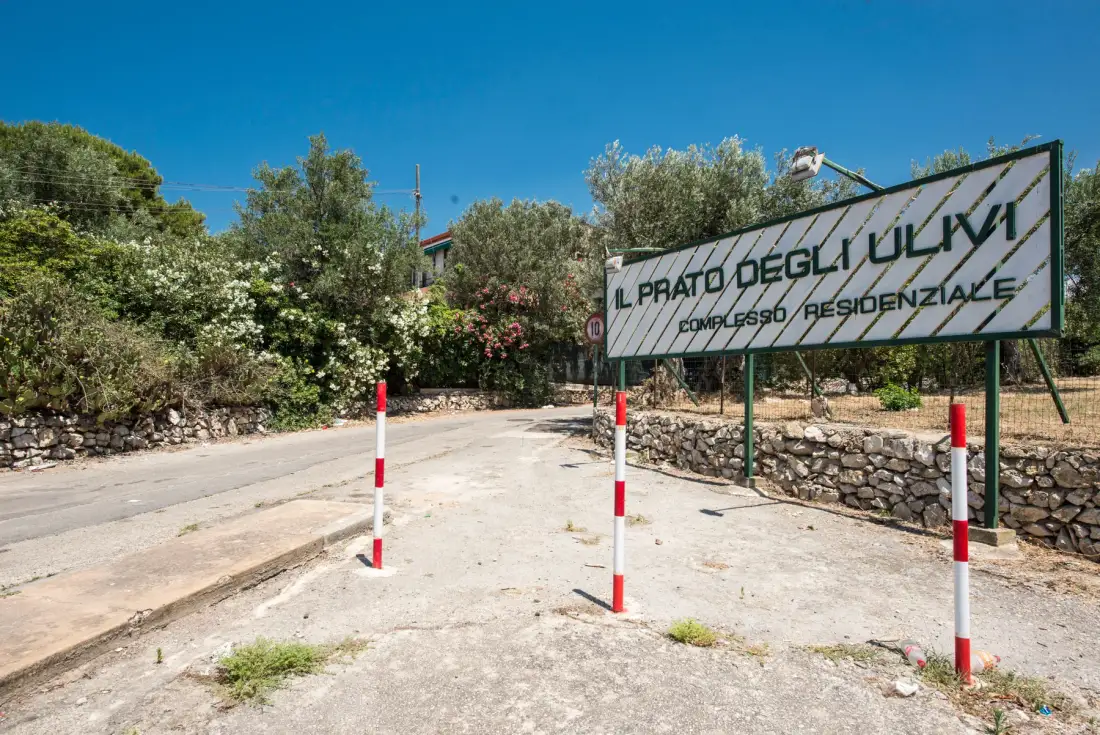 A paved entrance road passes a large sign beside stone walls and trees.