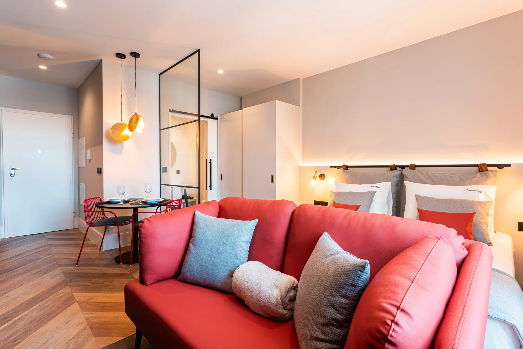 A studio room with a red sofa in front of a bed and dining table.