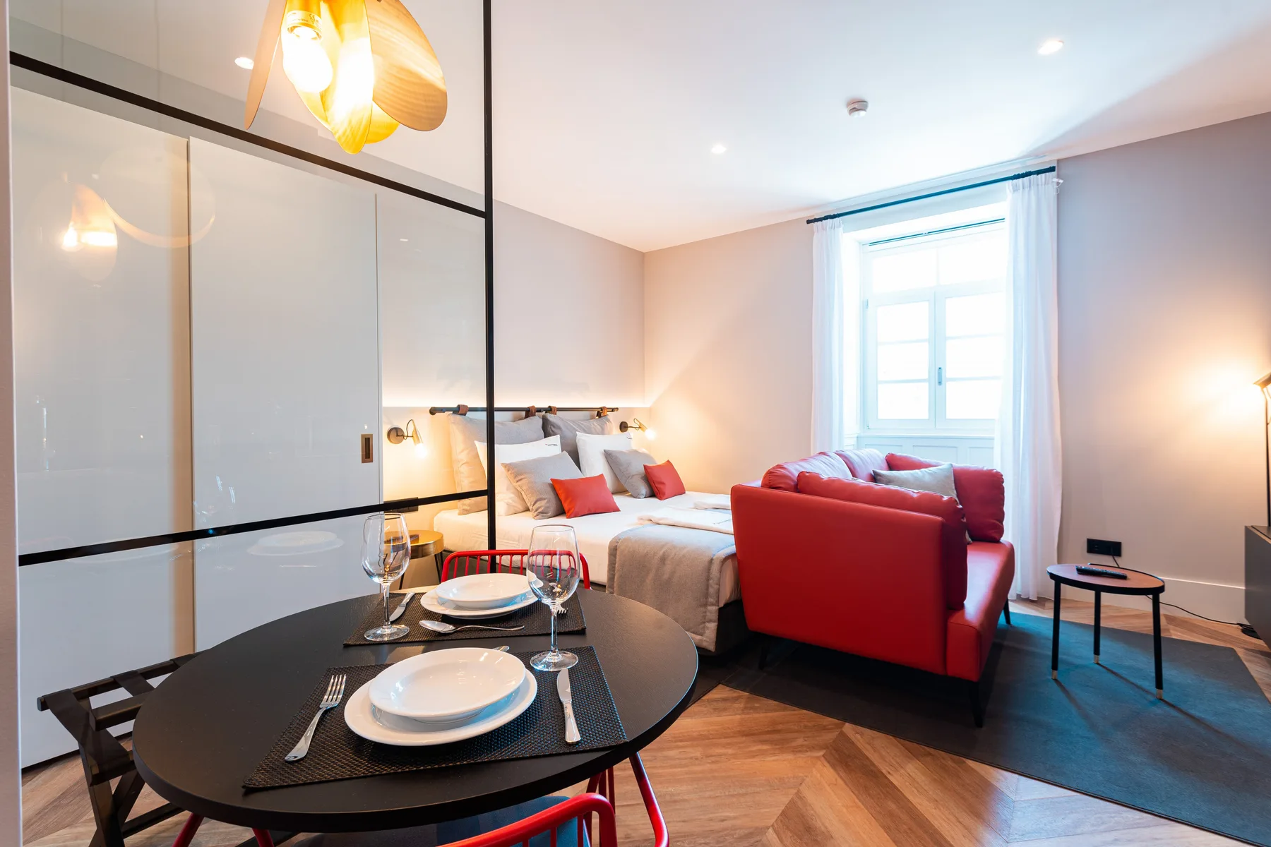 A studio room with a bed, red seating, and a dining table by the window.