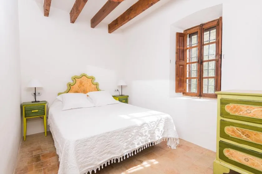 Un dormitorio luminoso con una cama blanca, mesitas de colores y una ventana de madera.