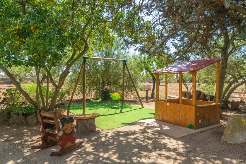 Una zona de juegos exterior a la sombra, con columpios y una pequeña casita de madera.