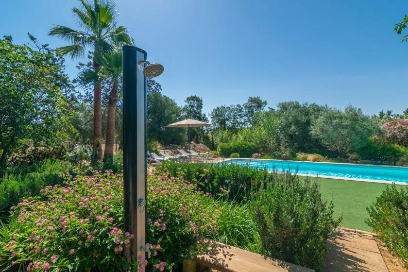 Una ducha exterior está junto a una piscina, rodeada de plantas verdes.