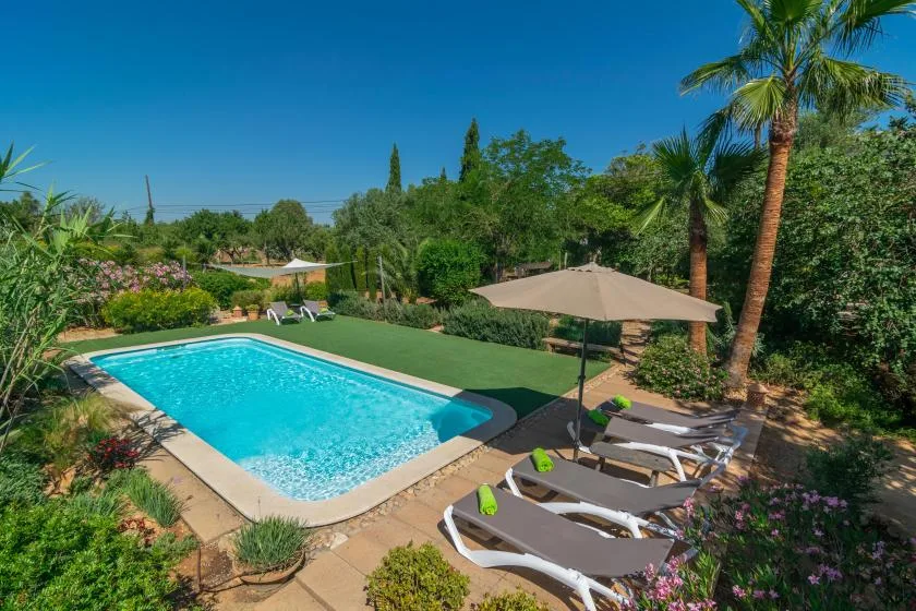 Una piscina al aire libre está junto a un jardín, con tumbonas bajo una sombrilla.