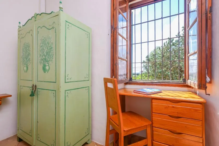 Un escritorio y una silla de madera están junto a una ventana grande, con un armario verde cerca.