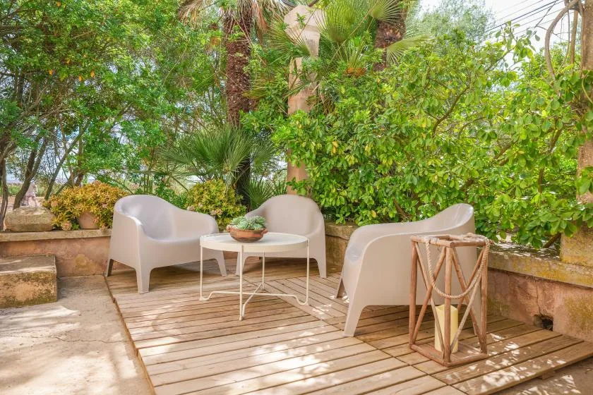 Una zona de estar al aire libre con asientos claros y una mesa pequeña, rodeada de plantas frondosas.