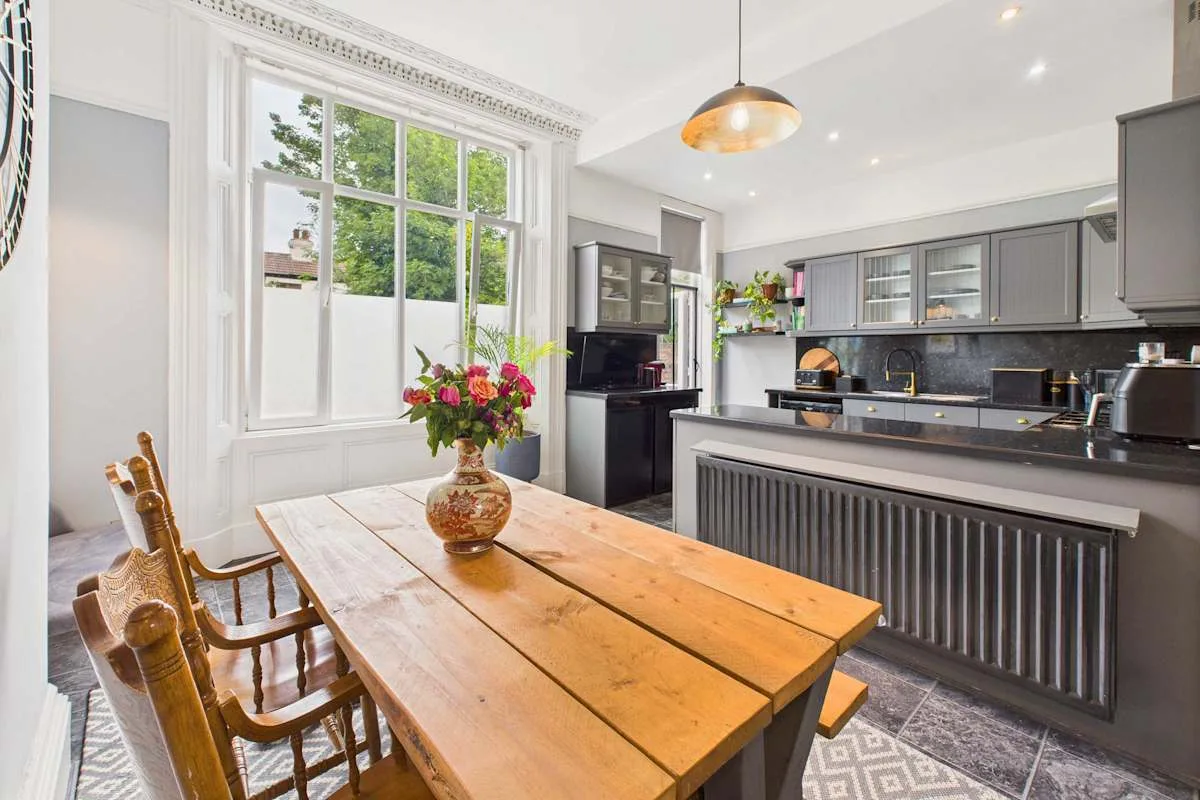 Una cocina luminosa con comedor, una mesa larga de madera y gabinetes oscuros.