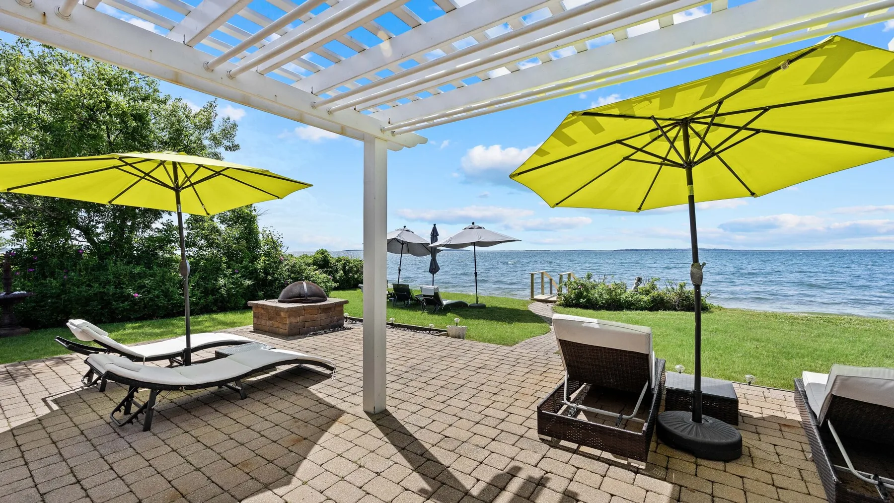 Un patio junto al lago tiene asientos bajo sombrillas amarillas y una pérgola blanca.