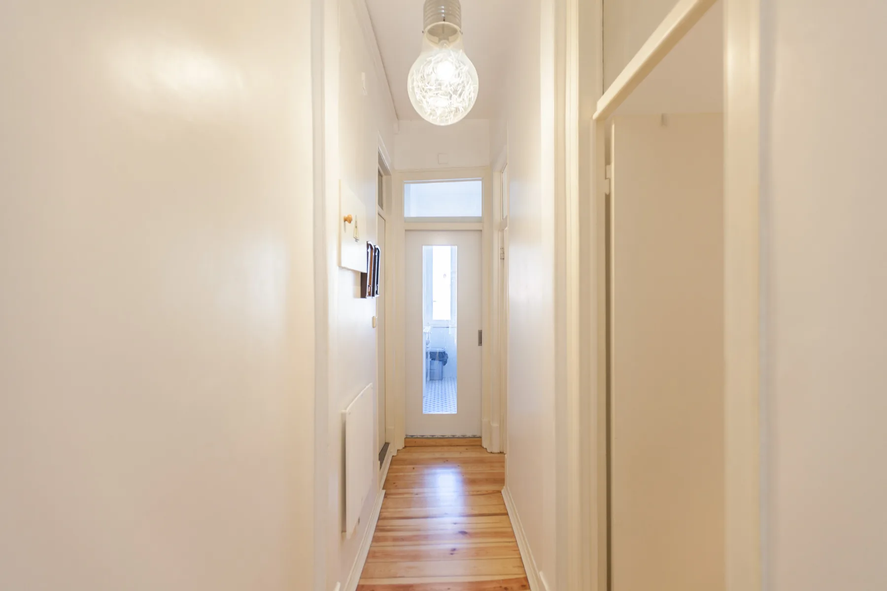 A narrow hallway with wood flooring leads to a bright glazed door.