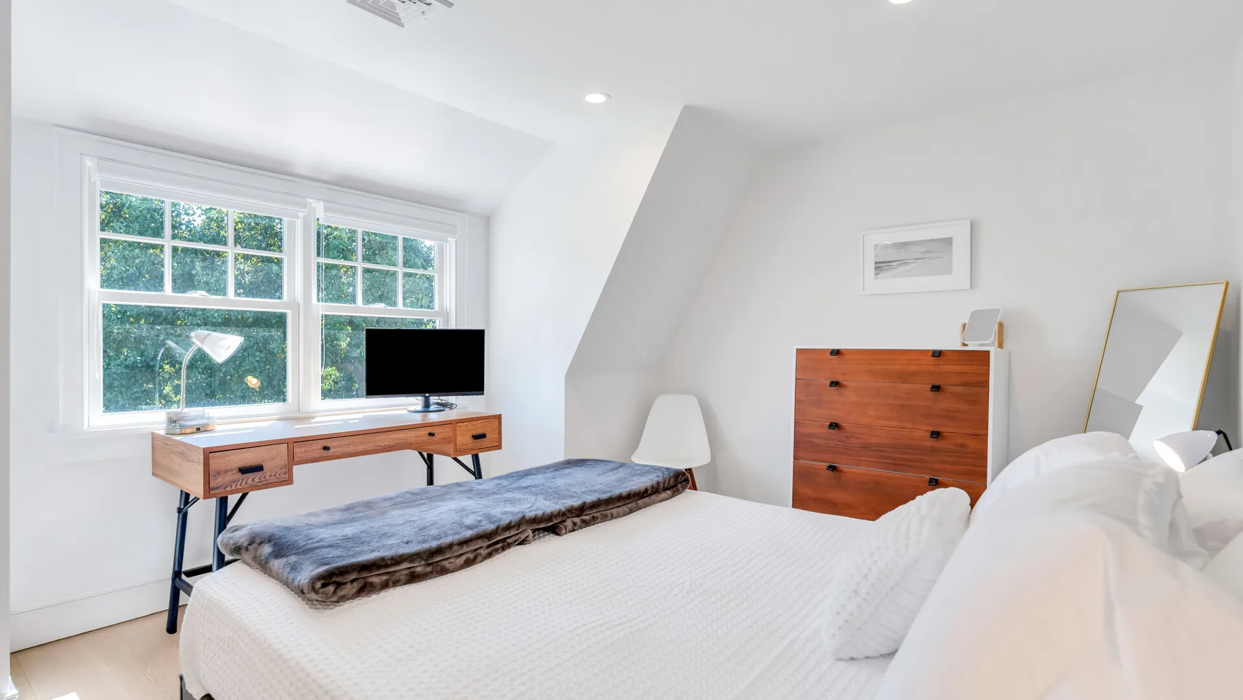 A bright bedroom with a bed, wooden desk, and dresser beside large windows.