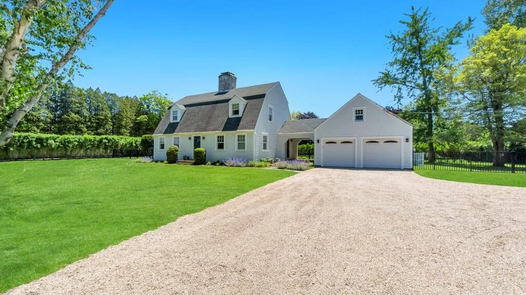 A white house and detached garage sit beside a broad gravel driveway and green lawn.