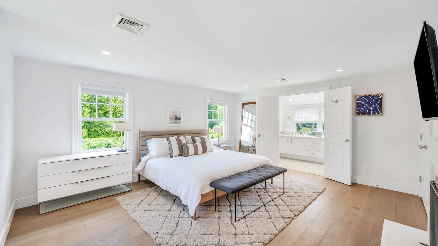 A bright bedroom with a bed, a bench, and windows overlooking greenery.
