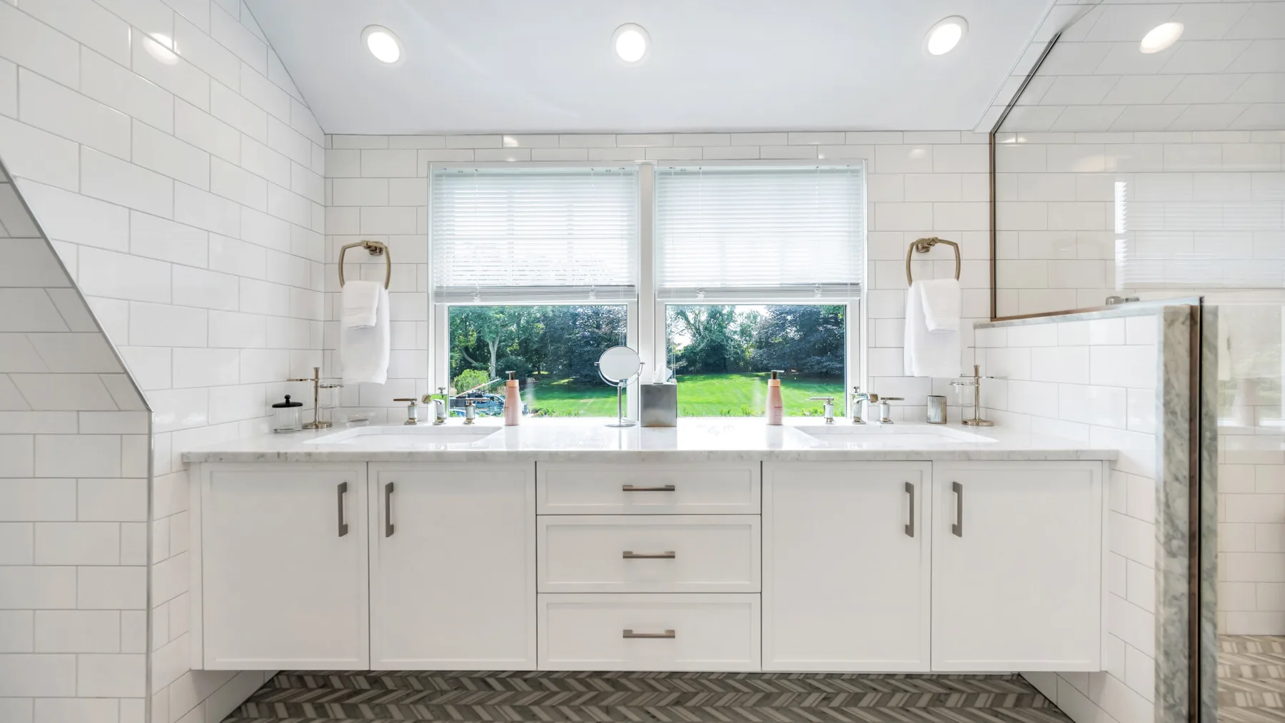 A bright bathroom vanity sits beneath two windows, with white tile walls and a glass shower.