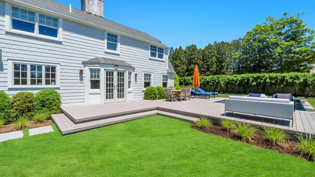 A sunny backyard patio beside a lawn, with outdoor seating and a swimming pool.